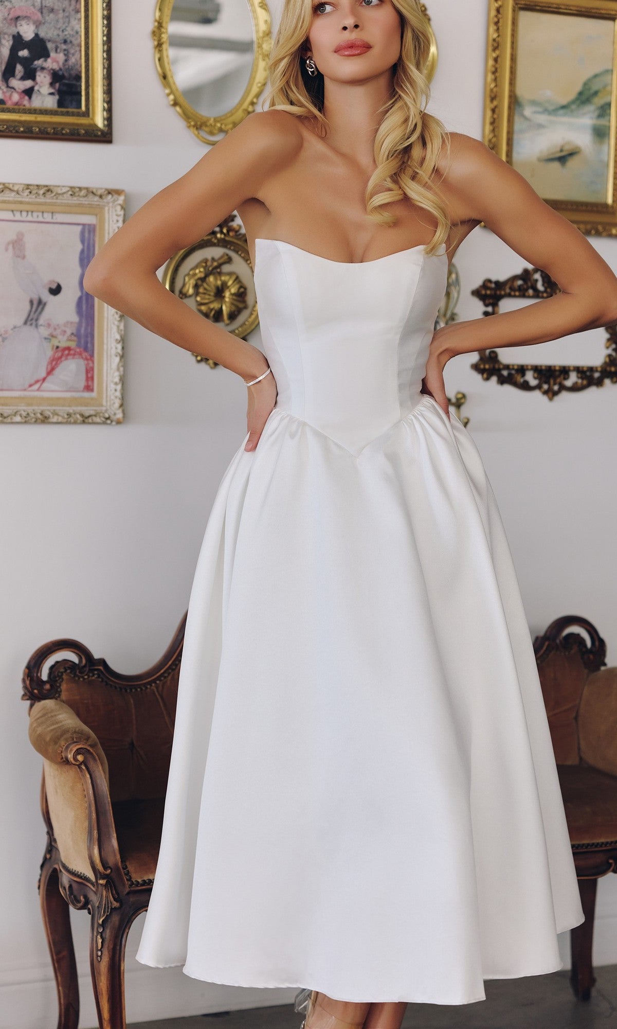 A woman stands indoors before a wall with framed art and an antique couch, wearing the Amoris T822W Off-White Midi Prom Dress featuring a strapless design and lace-up back.